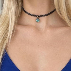 Peut inclure: Un collier ras-du-cou en dentelle noire avec un motif tissé délicat et un pendentif ovale unique orné d'une pierre irisée bleue et verte dans un cadre métallique, positionné au centre à la base du cou.