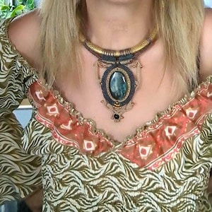 May include: A macrame necklace featuring a large oval dark green stone centerpiece surrounded by intricate dark blue and tan cord weaving. The necklace includes a textured gold-toned collar section and a small black bead accent at the bottom.