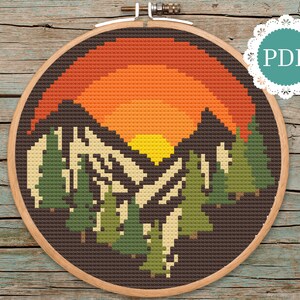 Könnte beinhalten: Kreuzstichmuster eines Sonnenuntergangs über einem Gebirge mit Bäumen im Vordergrund. Das Muster befindet sich in einem braunen Holzstickrahmen.