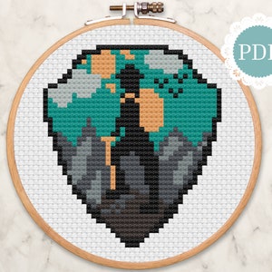 May include: Cross stitch pattern of a person standing on a mountain with a teal, orange, and grey colour scheme. The pattern is in a hoop and ready to be stitched.