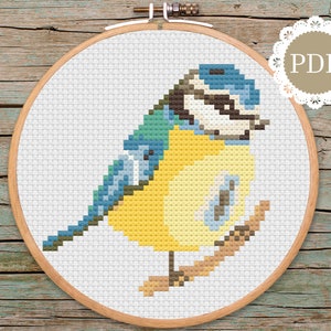 Może przedstawiać: Wzór haftu krzyżykowego przedstawiający sikorkę na gałęzi. Ptak jest żółty z niebieskimi skrzydłami i czarną głową. Wzór znajduje się w drewnianej obręczy do haftu.