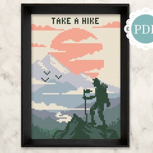 Op de afbeelding: Borduurpatroon met een wandelaar met een rugzak die op een bergtop staat met een zonsondergang op de achtergrond. De tekst "TAKE A HIKE" staat bovenaan het patroon.