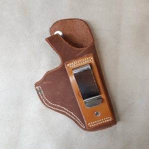May include: A brown leather holster with a metal clip. The holster has white stitching around the edges.