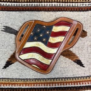 May include: A leather holster with an American flag design. The flag is red, white, and blue with a distressed look. The holster is brown and has stitching around the edges.