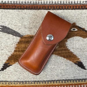 May include: A brown leather sheath with a snap closure. The sheath is designed to hold a small knife or tool.