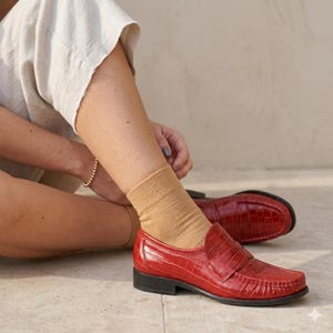 Op de afbeelding: Rode leren loafers met een krokodillenstructuur. De schoenen hebben een klassiek ontwerp met een lage hak en een zwarte zool. De loafers worden gecombineerd met beige sokken en een neutrale broek.