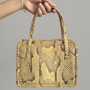 Op de afbeelding: Een rechthoekige handtas met een gestructureerd slangenleerpatroon in tinten beige, geel en donkerbruin. De tas heeft twee bijpassende handvatten en een gestructureerd frame, met een opvallende schubachtige textuur over de gehele buitenkant.