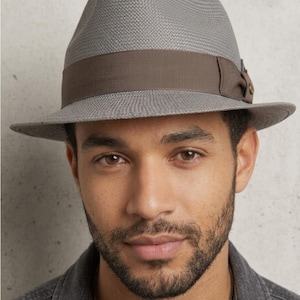 May include: A grey fedora hat with a textured, woven surface and a wide, dark brown grosgrain ribbon band. The hat features a pinched crown and a structured brim, with a small metallic detail on the side of the ribbon band.