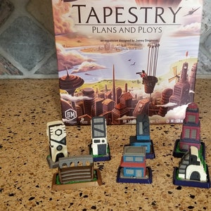 May include: A set of seven miniature buildings for the board game Tapestry: Plans and Ploys. The buildings are made of plastic and are painted in various colors, including white, gray, red, and blue. The buildings are arranged on a brown surface.