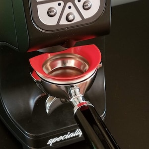 Puede incluir: Primer plano de un molinillo de café espresso negro con una cesta de portafiltro roja y un mango de portafiltro plateado. El molinillo tiene una pantalla digital y varios botones. El texto "specially" es visible en la base del molinillo.