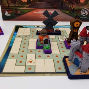 May include: A game board with a grid of squares and orange dots. There are several miniature buildings on the board, including a windmill, a clock tower, a pumpkin house, and a castle. The game board is made of cardboard and the buildings are made of plastic.