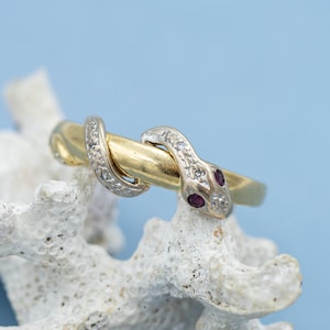 May include: A gold ring shaped like a snake with a silver-toned head and tail. The snake features small clear stones along its body and two small red stones for eyes. The ring is resting on a piece of white, textured, branch-like coral.