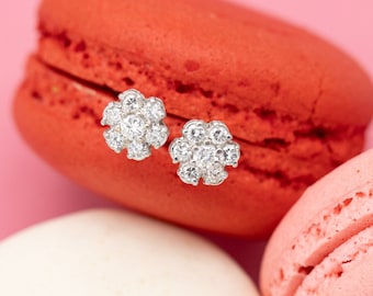 May include: A pair of flower-shaped stud earrings with clear gemstones, resting on a red macaron. The earrings are set against a pink background, with two other macarons in white and pink.