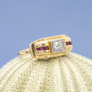 Peut inclure: Une bague en or avec une face rectangulaire ornée d'un diamant carré central et de deux rubis rectangulaires. La bague est posée sur une surface texturée blanc cassé sur fond bleu.