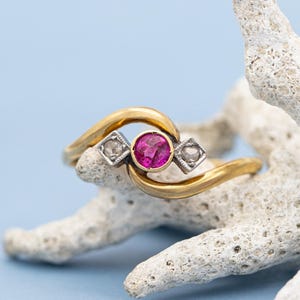 Pode incluir: Um anel de ouro com um rubi redondo central e dois diamantes laterais. O anel está colocado em um galho de coral branco sobre um fundo azul claro. O design do anel apresenta uma faixa de ouro em espiral.