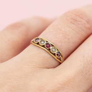Puede incluir: Un anillo de oro con una fila de piedras preciosas rojas y blancas alternas. El anillo se lleva en un dedo sobre un fondo rosa suave. Las piedras preciosas están engastadas en una banda curva y calada.