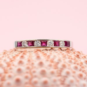 Puede incluir: Un anillo de plata con gemas rojas cuadradas y gemas claras redondas alternadas. El anillo está sobre un fondo rosa y descansa sobre una superficie texturizada de color rosa claro.