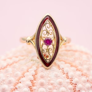 May include: A gold ring with a ruby gemstone. The ring features a maroon-colored enamel design with an ornate filigree pattern surrounding the ruby. The ring is set on a pink, textured surface.