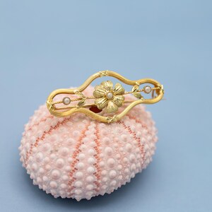 Puede incluir: Broche dorado con dise&ntilde;o floral, perla central y dos perlas m&aacute;s peque&ntilde;as. La forma de la broche es curva, con detalles florales y hojas. Descansa sobre una superficie rosa texturizada sobre un fondo azul claro. Joyer&iacute;a vintage.
