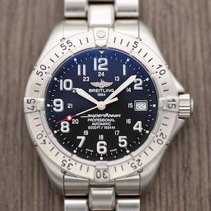 May include: A stainless steel Breitling Superocean Professional automatic wristwatch. The watch has a black dial with white numerals and hands, a date window, and a rotating bezel. The watch is on a stainless steel bracelet.
