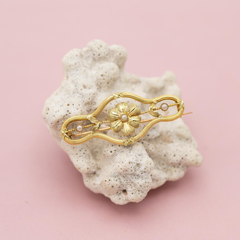 Puede incluir: Broche dorado con dise&ntilde;o floral y dos peque&ntilde;as perlas. El broche presenta una flor central con un centro de perla, engastada en un marco calado. El broche se muestra sobre una superficie blanca texturizada sobre un fondo rosa.