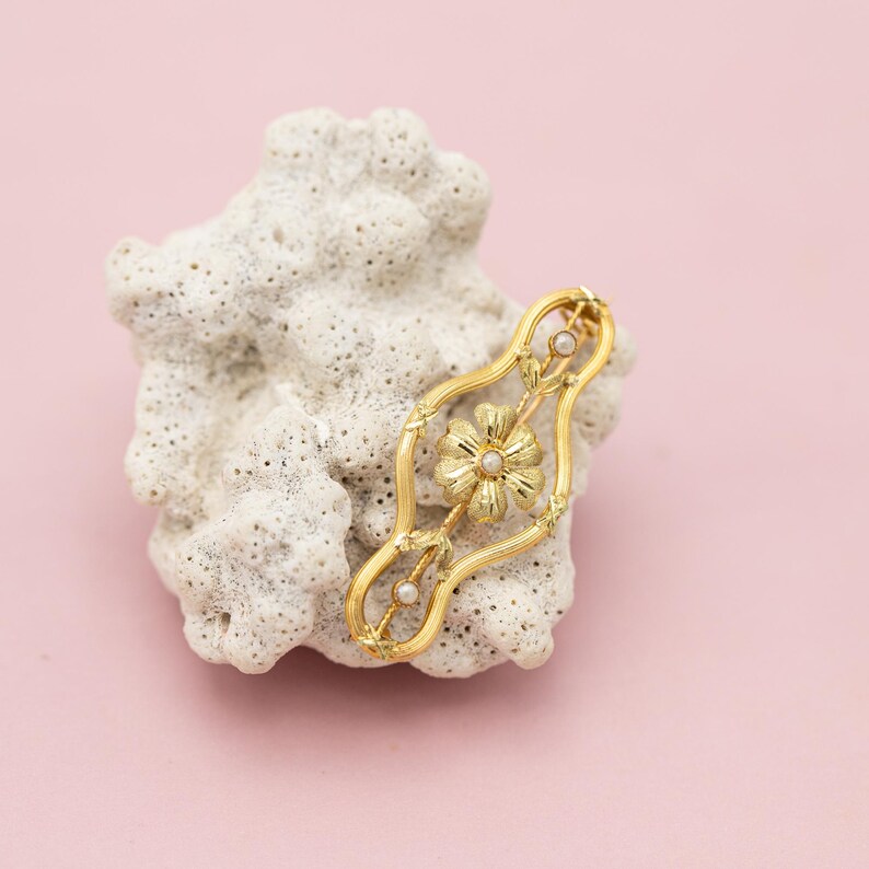 Puede incluir: Broche dorado con dise&ntilde;o floral y peque&ntilde;os detalles de perlas. La broche calada se encuentra sobre un fondo blanco texturizado con un fondo rosa. Joya de estilo vintage.