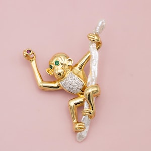 Op de afbeelding: Een goudkleurige apenbroche met een wit touw. De aap heeft groene ogen en een robijn in zijn hand. De borst van de aap is bezet met kleine diamanten. De achtergrond is effen lichtroze.