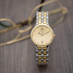 Puede incluir: Reloj Omega bicolor en oro y plata. La esfera es de color oro claro, con visualización de la fecha. Tiene un bisel dorado y una pulsera de eslabones plateados y dorados. Fondo borroso con gafas.