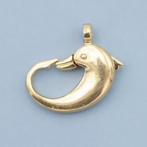 May include: A gold-colored dolphin-shaped clasp. The clasp features a curved body, a beak-like closure, and a small loop at the top. The dolphin has a smooth, polished surface and a small fin detail. The background is a solid light blue.