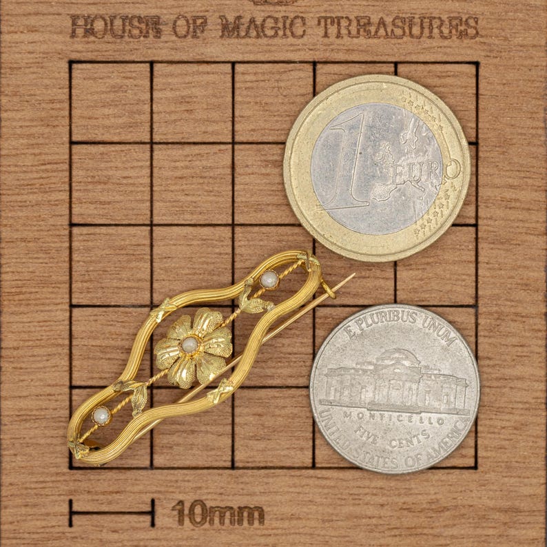 Puede incluir: Broche dorado con dise&ntilde;o floral y peque&ntilde;as perlas, sobre una cuadr&iacute;cula de madera. Una moneda de euro y un n&iacute;quel estadounidense se usan para comparar el tama&ntilde;o. El texto "HOUSE OF MAGIC TREASURES" es visible. La longitud de la broche es de unos 3 cm.