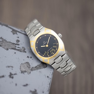May include: An Omega Seamaster watch with a two-tone silver and gold stainless steel link bracelet. The watch features a round black analogue dial with gold hands, gold hour markers, and a digital display at the bottom showing the numbers 22.09.
