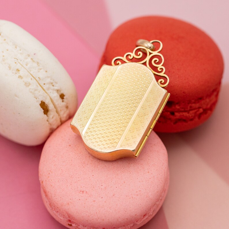 Może przedstawiać: Złoty medalion z ozdobnym wzorem leży na dw&oacute;ch r&oacute;żowych makaronach. Medalion ma fakturowaną powierzchnię i otwierane na zawiasach.
