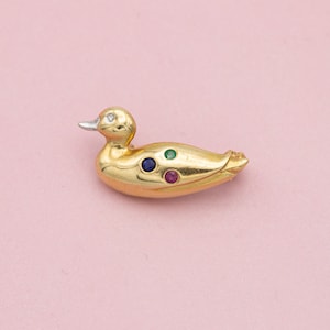 Puede incluir: Broche de pato dorado con un ojo de diamante y un pico plateado. El cuerpo del pato presenta tres piedras preciosas engastadas: azul, verde y roja. El broche está sobre un fondo rosa suave.
