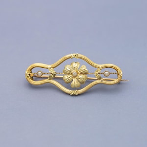 Puede incluir: Broche dorado con un dise&ntilde;o floral central y dos peque&ntilde;as perlas engastadas en un marco curvo decorativo. El broche tiene un cierre de alfiler. El dise&ntilde;o es sim&eacute;trico, con una flor en el centro y una perla a cada lado.
