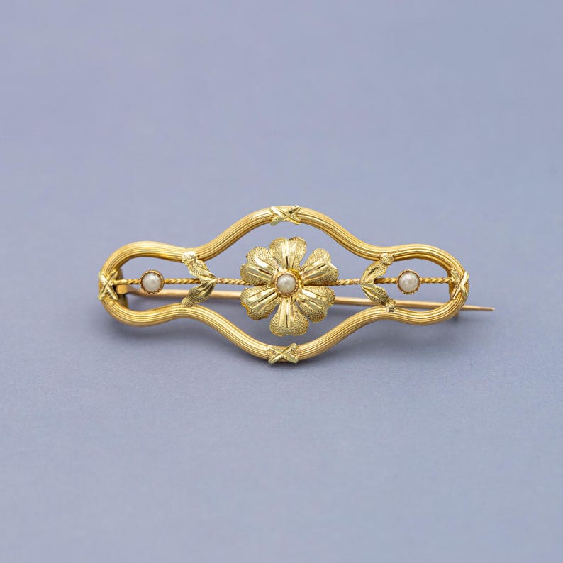 Puede incluir: Broche dorado con un dise&ntilde;o floral central y dos peque&ntilde;as perlas engastadas en un marco curvo decorativo. El broche tiene un cierre de alfiler. El dise&ntilde;o es sim&eacute;trico, con una flor en el centro y una perla a cada lado.