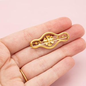 Puede incluir: Un broche dorado con un dise&ntilde;o floral. El broche presenta una flor central y dos peque&ntilde;os detalles florales, todo dentro de un marco decorativo y alargado. El alfiler se sostiene en una mano sobre un fondo rosa.