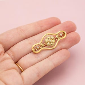 Puede incluir: Un broche de barra dorado con un dise&ntilde;o floral central y un peque&ntilde;o centro de perla. El broche tiene una forma alargada con elementos decorativos y dos peque&ntilde;as perlas. La joya se sostiene en una mano sobre un fondo rosa.