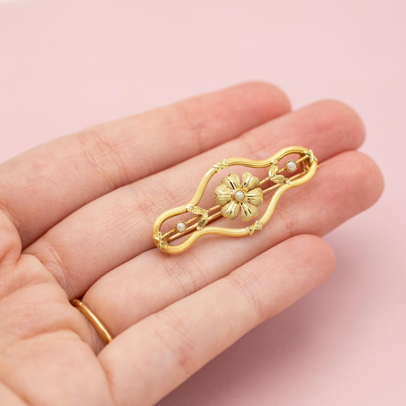 Puede incluir: Un broche de barra dorado con un dise&ntilde;o floral central y un peque&ntilde;o centro de perla. El broche tiene una forma alargada con elementos decorativos y dos peque&ntilde;as perlas. La joya se sostiene en una mano sobre un fondo rosa.