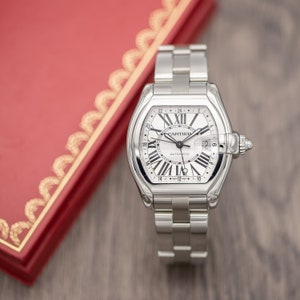 Puede incluir: Un reloj de pulsera plateado con esfera blanca y números romanos negros. El reloj tiene una pantalla de fecha y está colocado sobre una caja roja con ribete dorado. La correa del reloj es de eslabones plateados.