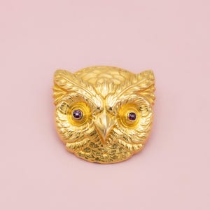 May include: A gold-colored owl brooch with purple gemstone eyes. The brooch is highly detailed, with textured feathers and a prominent beak. The owl is centered against a soft pink background, creating a striking contrast.