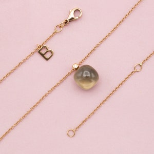 Puede incluir: Un collar dorado con un colgante de gema cuadrada translúcida gris-amarilla. El collar presenta una delicada cadena, un pequeño acento de diamante y un cierre de mosquetón. Un dije de oro con la letra "B" cuelga.