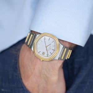 Puede incluir: Un reloj de pulsera dorado y plateado con una esfera de color claro y una pantalla de fecha en la parte inferior. El reloj tiene un bisel dorado y una correa plateada. El reloj está en una muñeca, con una manga de camisa azul claro en el fondo.