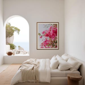 May include: A white bed with a white linen duvet cover and a beige throw blanket. The bed is in a white room with a large arched window overlooking a view of the ocean and a rocky coastline. A framed print of pink flowers hangs on the wall above the bed.