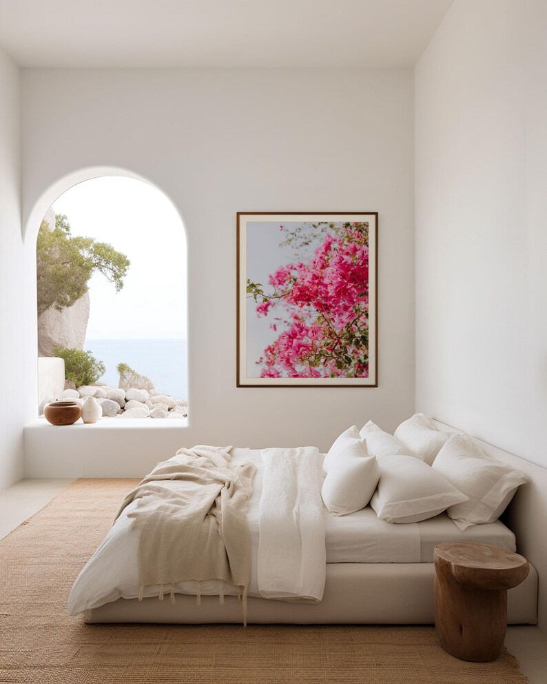 May include: A white bed with a white linen duvet cover and a beige throw blanket. The bed is in a white room with a large arched window overlooking a view of the ocean and a rocky coastline. A framed print of pink flowers hangs on the wall above the bed.