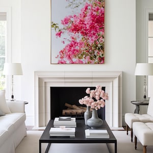 May include: A white living room with a fireplace and a black coffee table. The fireplace has a black hearth and a white mantel. The coffee table has a black frame and a wooden top. There are books and a vase of pink flowers on the table. A framed print of pink flowers hangs above the fireplace.