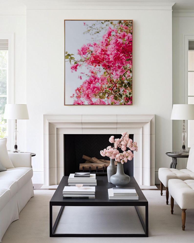 May include: A white living room with a fireplace and a black coffee table. The fireplace has a black hearth and a white mantel. The coffee table has a black frame and a wooden top. There are books and a vase of pink flowers on the table. A framed print of pink flowers hangs above the fireplace.