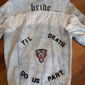 May include: A light blue acid-wash denim jacket featuring white fringe across the back yoke. The back displays the black text "bride" above the fringe, and "TIL DEATH DO US PART" below it, surrounding a central panther head patch and silver stars.
