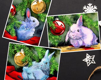 Pode incluir: Três aquarelas de coelhos debaixo de uma árvore de Natal com ornamentos. O primeiro coelho é azul, o segundo é roxo e o terceiro é azul. Os ornamentos são dourados, vermelhos e dourados.