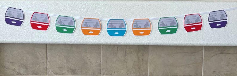 May include: A colourful banner with a string of paper cutouts shaped like a train car. The train cars are in shades of purple, red, green, orange, blue, and green. Each car has a window with a silhouette of people inside.