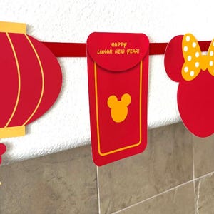 Puede incluir: Bandera roja y amarilla del A&ntilde;o Nuevo Chino con linternas rojas, sobres rojos y una cabeza de Mickey Mouse con un lazo amarillo. Las linternas tienen el a&ntilde;o 2025 impreso en ellas. Los sobres rojos tienen el texto "&iexcl;Feliz A&ntilde;o Nuevo Lunar!" impreso en ellos.